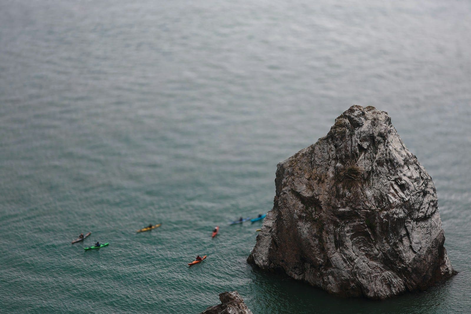 Seen from above, a group of sea kayakers paddles past a large sea stack in gray-green ocean water