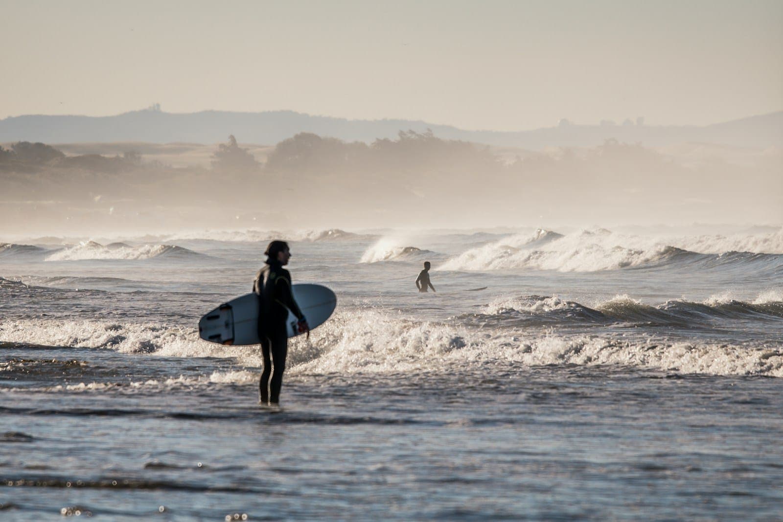 A surfer in a wetsuit stands knee-deep holding a white surfboard, with small soft waves behind on an overcast day