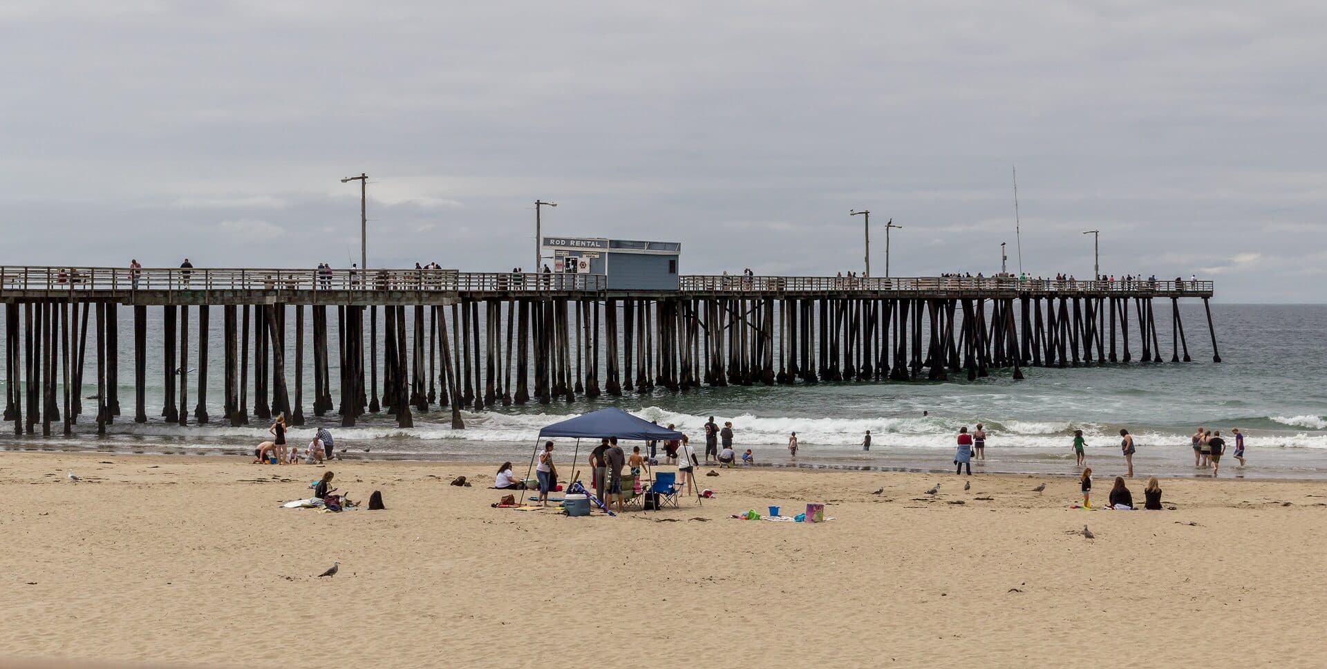 Pismo Beach Pier on an overcast day, with small surf breaking under the pilings and families on the sand
