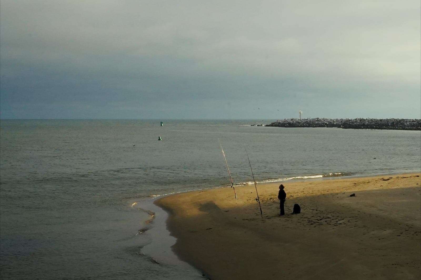 A lone angler with rods in sand spikes on the beach at Morro Bay, the harbor-mouth breakwater behind, under cloudy skies