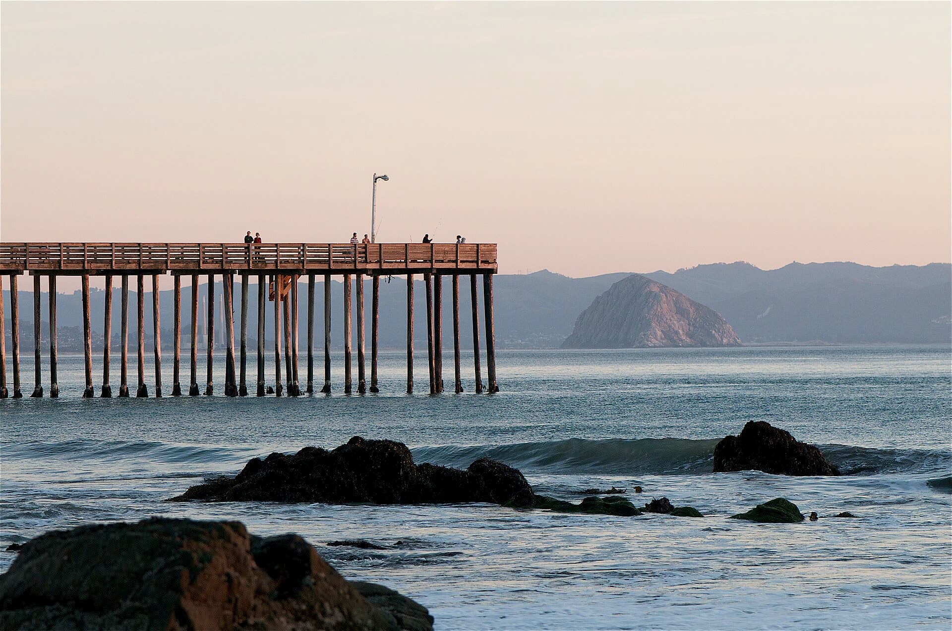 The Cayucos Pier on wooden pilings over calm water at dusk, with dark rocks in the foreground and Morro Rock across Estero Bay
