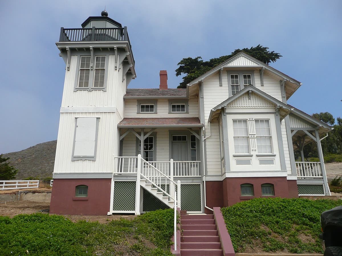 The white Victorian Point San Luis Lighthouse, with its square tower and red-trimmed base, on a green lawn