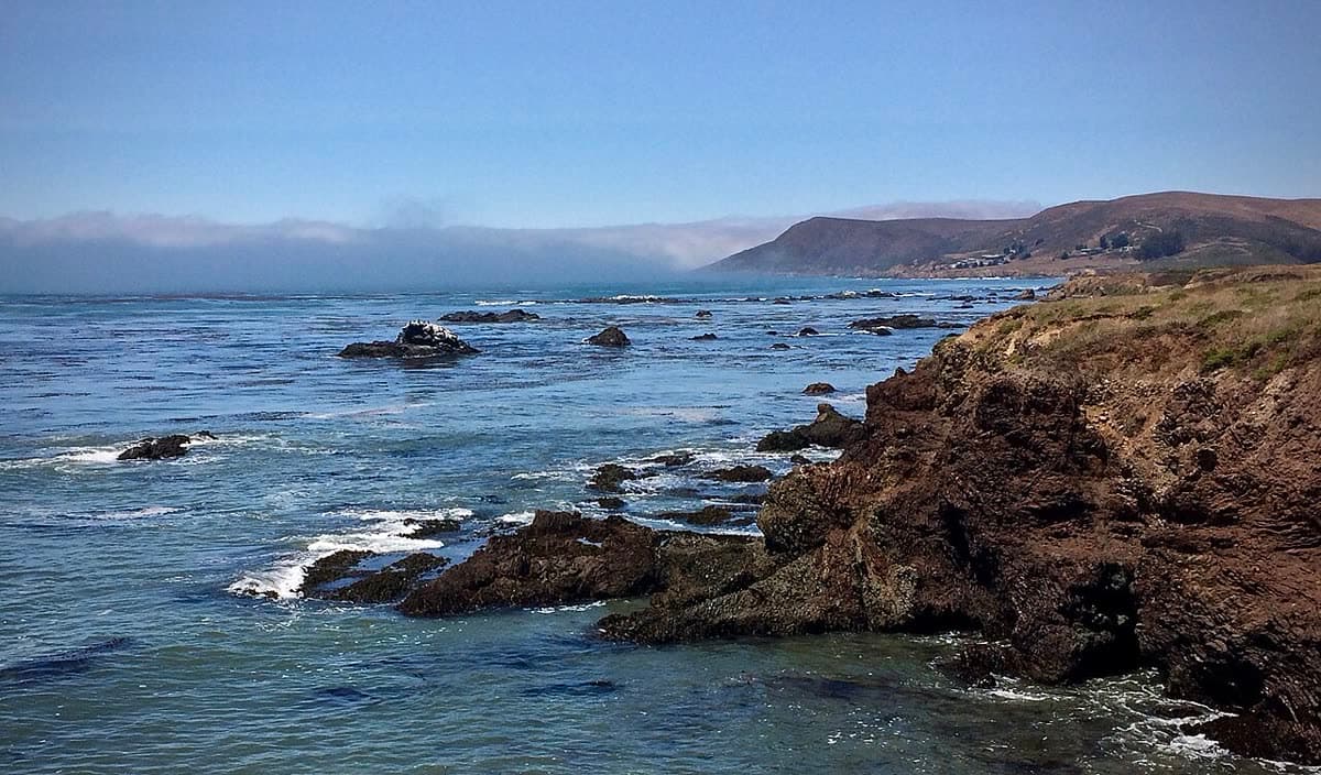 Dark rocky headland and offshore rocks at Estero Bluffs State Park, with a fog bank rolling in over the hills behind